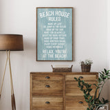 A Beach House Rules Sign in painted wood (Tide) hangs above a wooden dresser with plants, bringing coastal wall art charm to this cozy room.