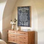 a wooden dresser with a framed sign that reads "BEACH HOUSE RULES" hanging above it. The dresser has a vase of white flowers and a stack of books on top.