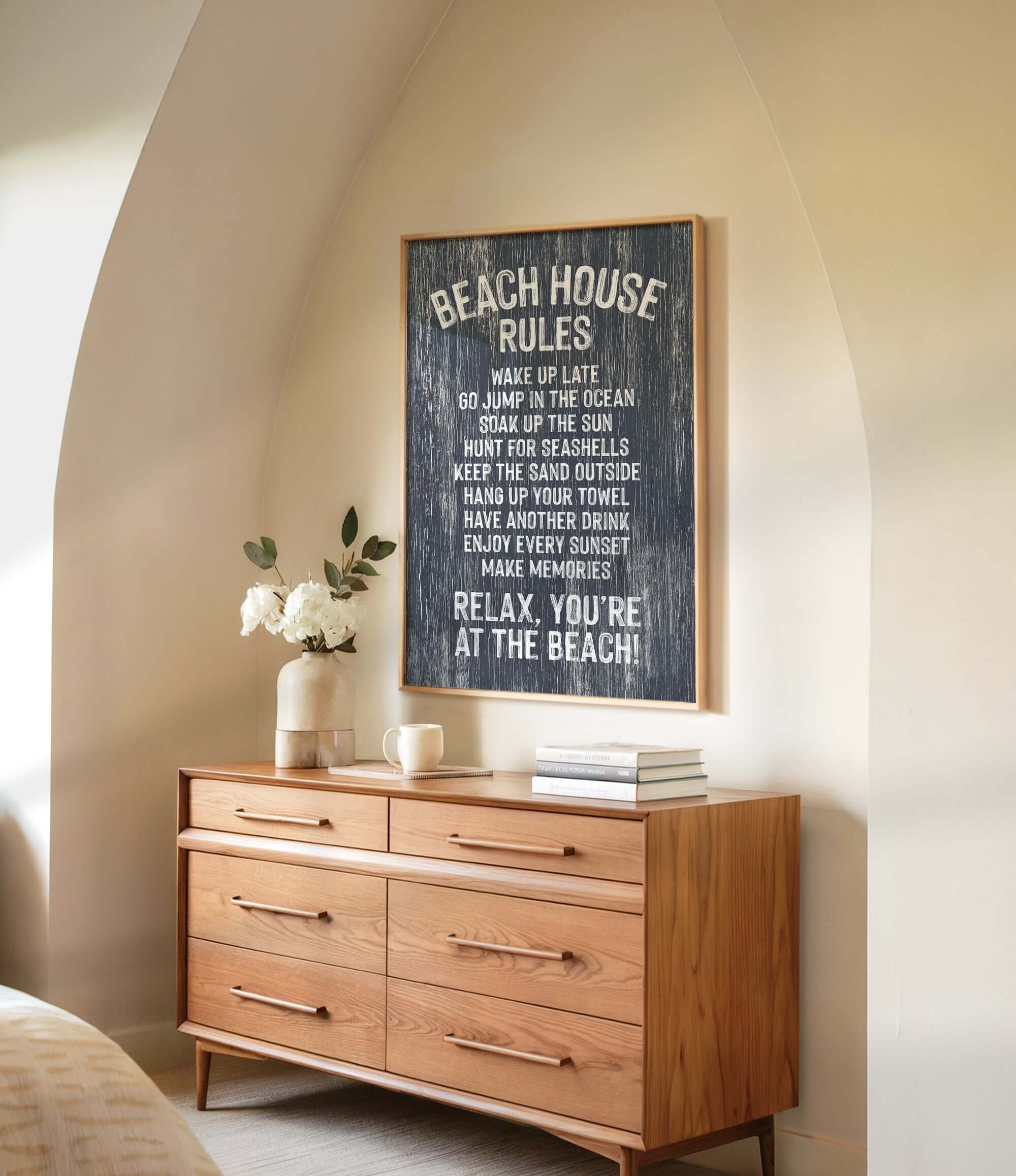 a wooden dresser with a framed sign that reads "BEACH HOUSE RULES" hanging above it. The dresser has a vase of white flowers and a stack of books on top.