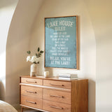 a wooden dresser with a framed sign that reads "LAKESHOUSE RULES" hanging on the wall above it. The dresser has a vase of white flowers and a stack of books on top.