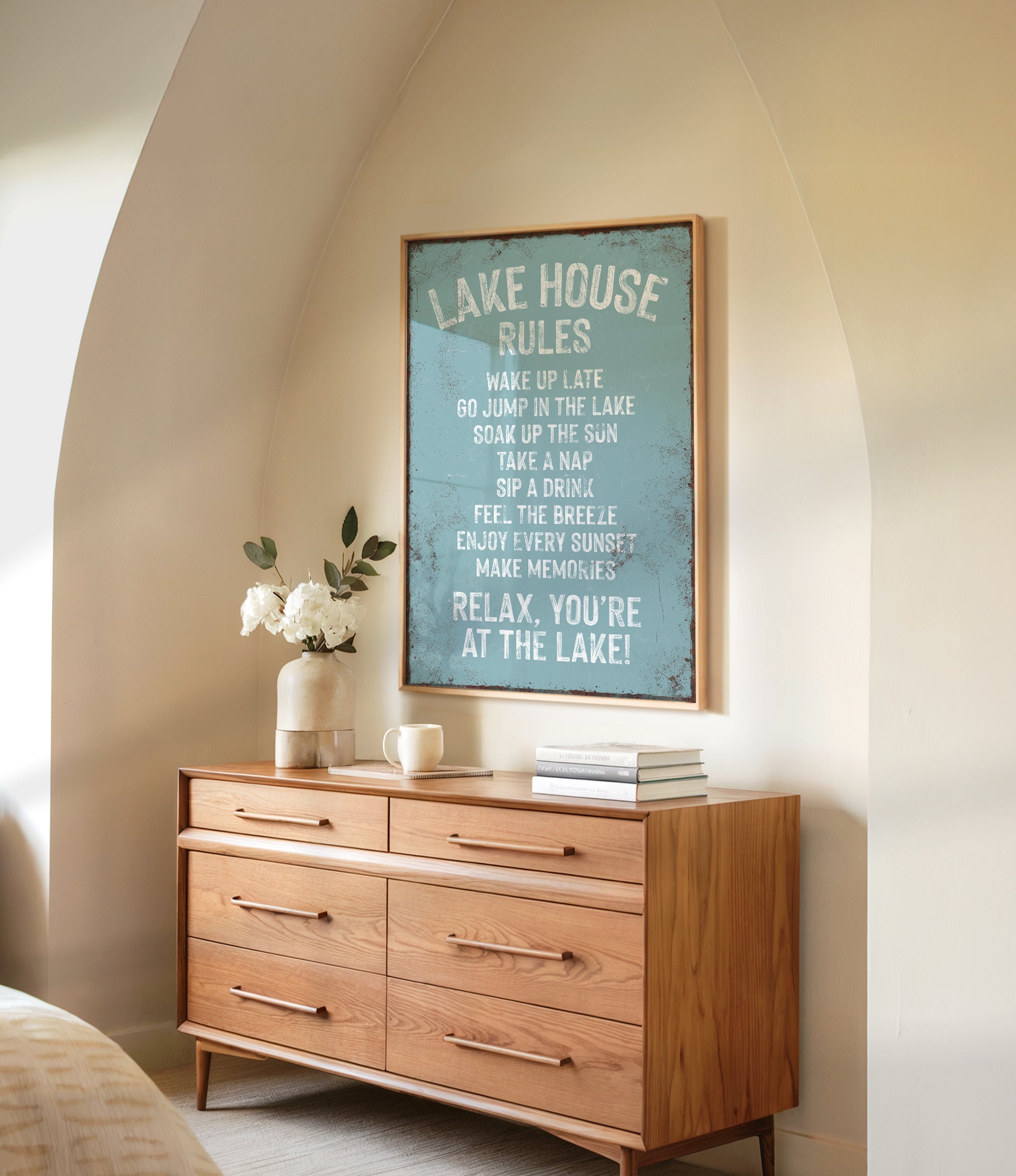 a wooden dresser with a framed sign that reads "LAKESHOUSE RULES" hanging on the wall above it. The dresser has a vase of white flowers and a stack of books on top.