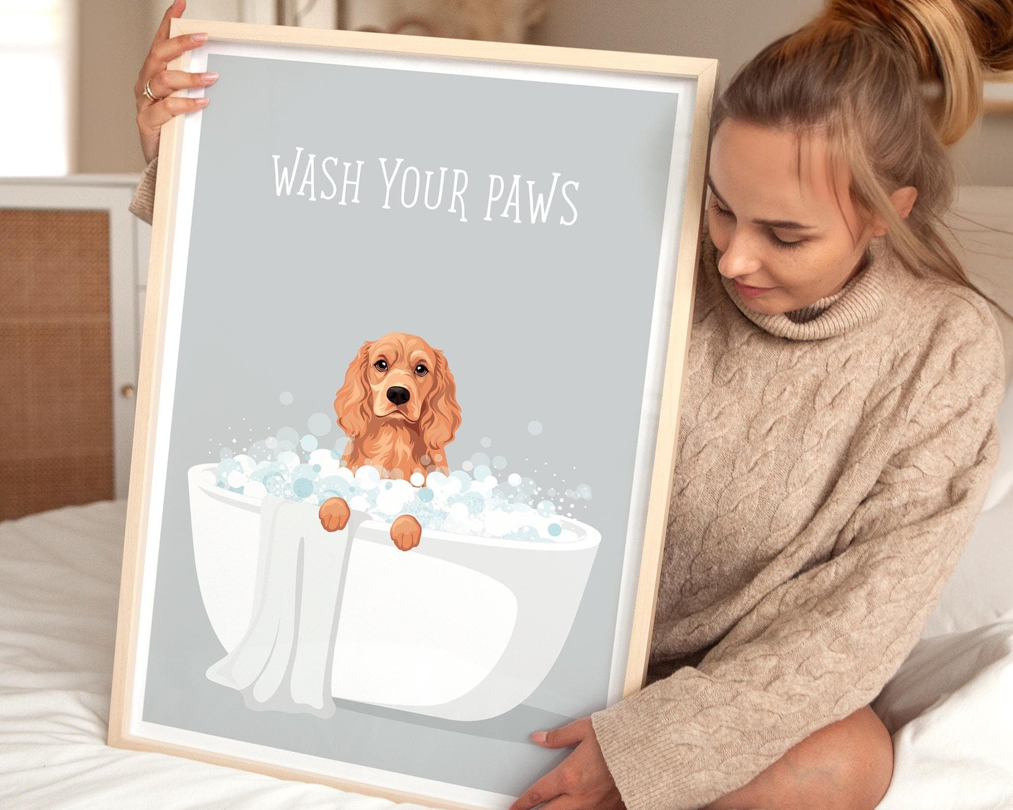 A woman is sitting on a bed holding a framed poster with a dog sitting in a bathtub filled with bubbles.