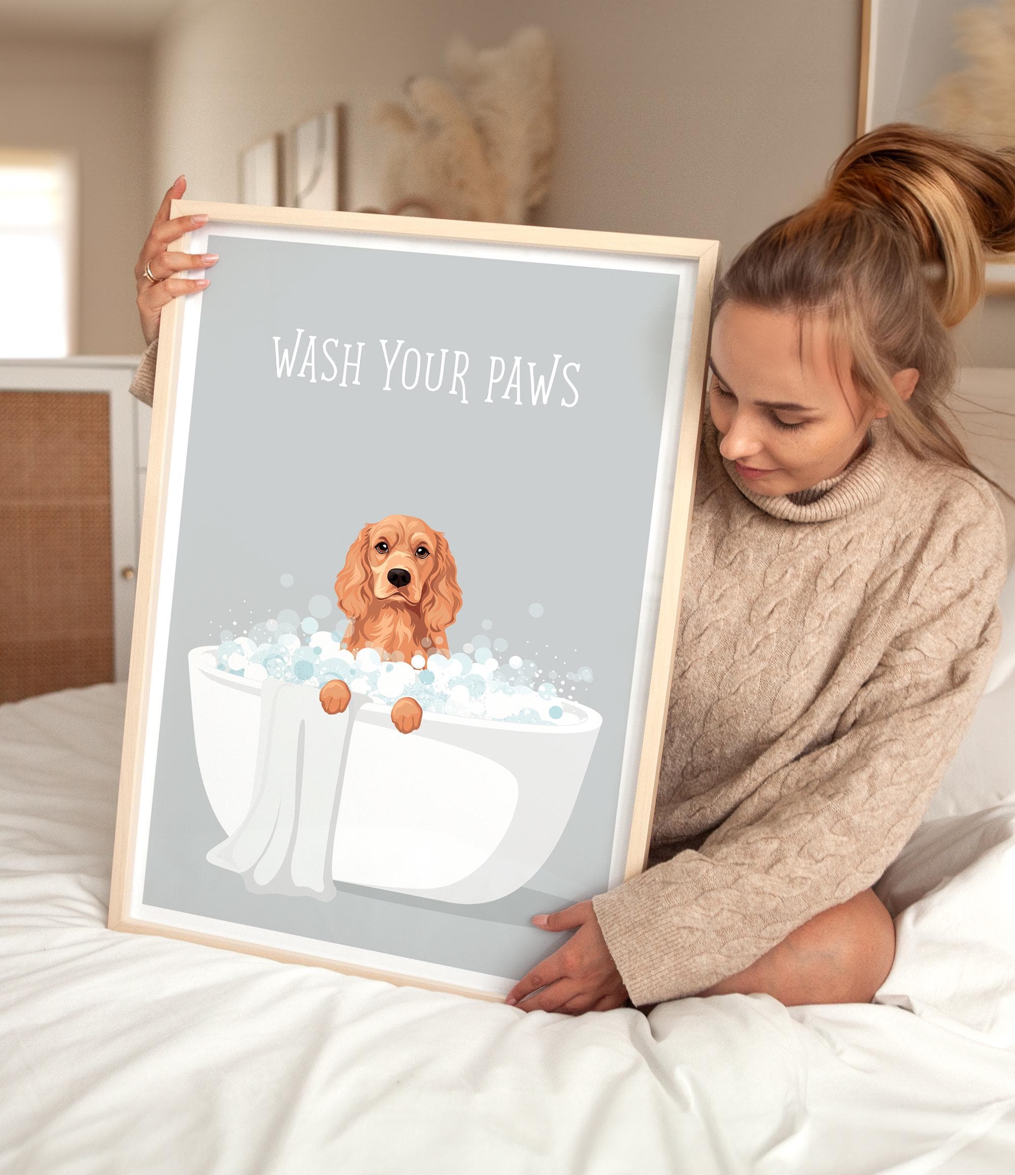 A woman is sitting on a bed holding a framed poster with a dog sitting in a bathtub filled with bubbles.