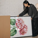 A woman is standing next to a large framed painting of a pink flower, examining it closely.