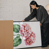 A woman is standing next to a large framed painting of a pink flower, examining it closely.