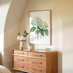 a wooden dresser with a framed painting of green hydrangeas hanging on the wall above it. The dresser has six drawers and a vase with white flowers on top.