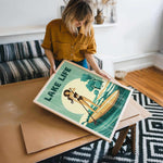 A woman is holding a framed poster with a beach scene and a woman on a surfboard.