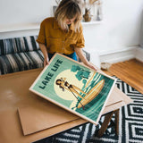 A woman is holding a framed poster with a beach scene and a woman on a surfboard.