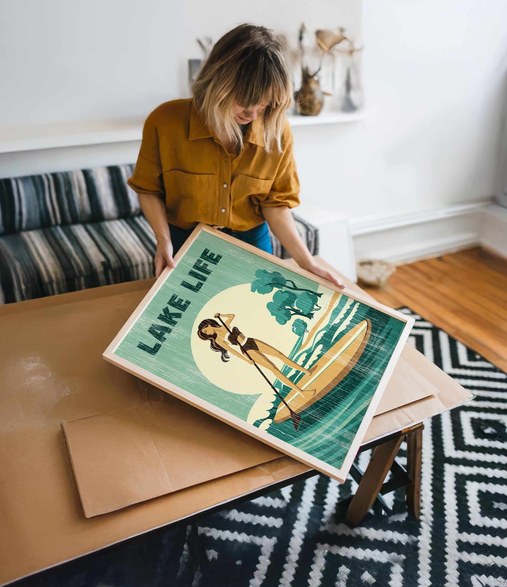A woman is holding a framed poster with a beach scene and a woman on a surfboard.