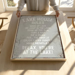 A person is holding up a framed sign with a list of rules for a lake house, including activities such as waking up late, jumping in the lake, taking a nap, soaking up the sun, sipping a drink, feeling the breeze, enjoying every sunset, and relaxing.