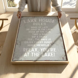 A person is holding up a framed sign with a list of rules for a lake house, including activities such as waking up late, jumping in the lake, taking a nap, soaking up the sun, sipping a drink, feeling the breeze, enjoying every sunset, and relaxing.