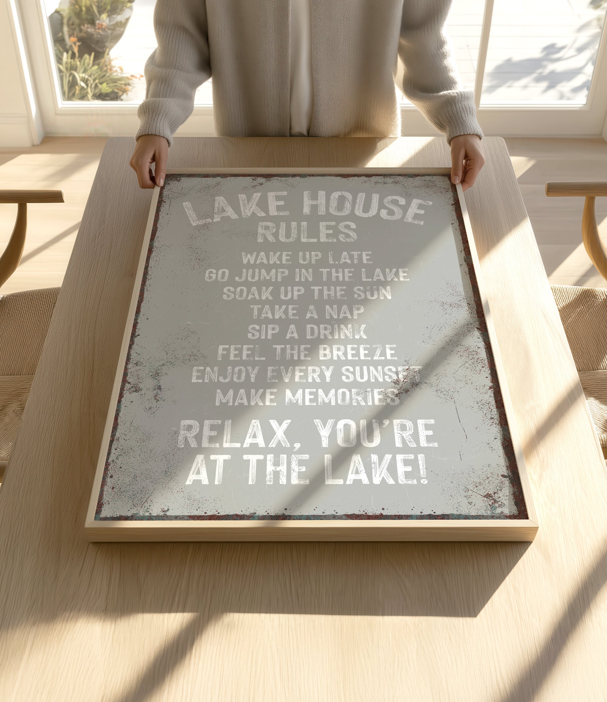 A person is holding up a framed sign with a list of rules for a lake house, including activities such as waking up late, jumping in the lake, taking a nap, soaking up the sun, sipping a drink, feeling the breeze, enjoying every sunset, and relaxing.