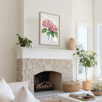 a cozy living room with a stone fireplace, a framed watercolor painting of pink hydrangeas hanging above it, and a white sofa. The room is decorated with potted plants and a woven basket, creating a warm and inviting atmosphere.