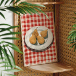A framed painting of three pears on a red and white checkered tablecloth is displayed on a wooden shelf, with a woven plant in the foreground.