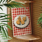 A framed painting of three pears on a red and white checkered tablecloth is displayed on a wooden shelf, with a woven plant in the foreground.