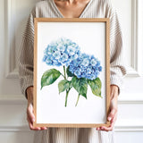 A woman is holding a framed watercolor painting of two blue hydrangea flowers.