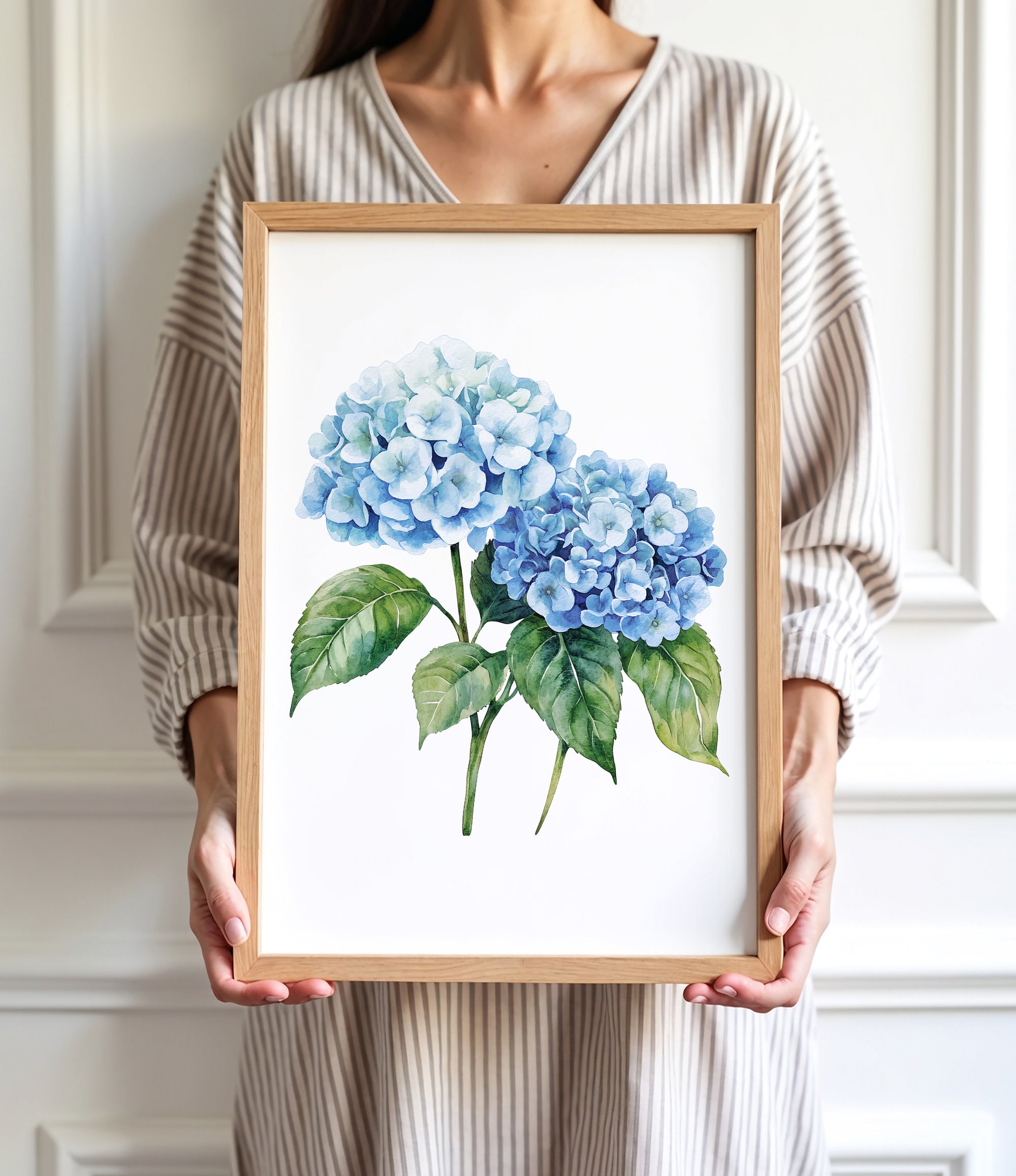 A woman is holding a framed watercolor painting of two blue hydrangea flowers.