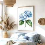 a serene bedroom scene with a large framed print of a blue hydrangea flower hanging on the wall above a bed with blue and white bedding. The room also features a woven basket, a potted plant, and a wooden nightstand with a vase.