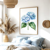 a serene bedroom scene with a large framed print of a blue hydrangea flower hanging on the wall above a bed with blue and white bedding. The room also features a woven basket, a potted plant, and a wooden nightstand with a vase.