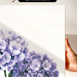 a hand holding a white canvas print of a watercolor painting of purple hydrangeas.