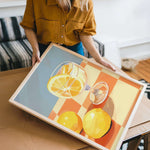 A woman is holding a framed painting of a glass of wine and two lemons on a table.