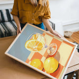 A woman is holding a framed painting of a glass of wine and two lemons on a table.