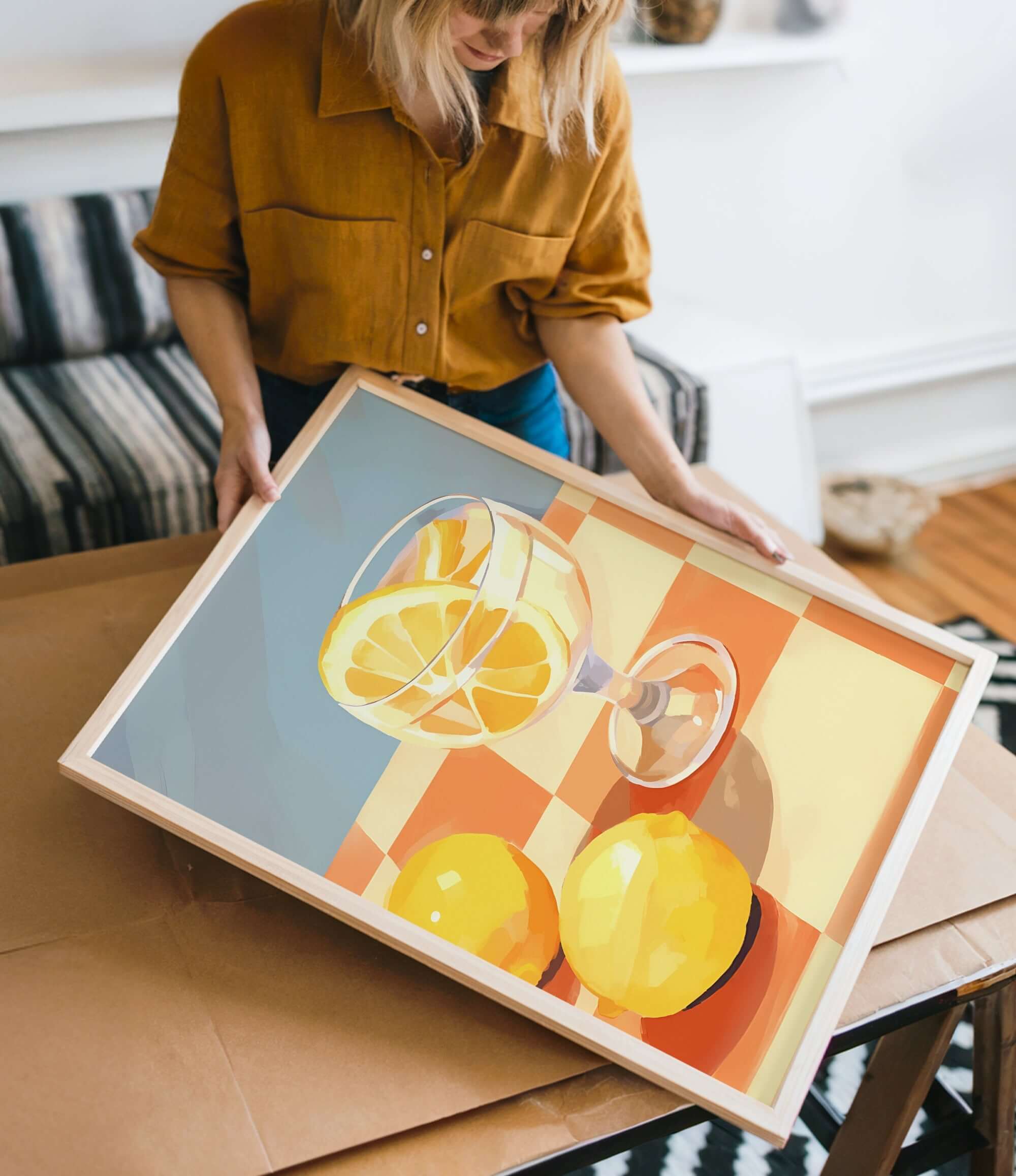 A woman is holding a framed painting of a glass of wine and two lemons on a table.