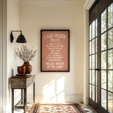 a hallway with a framed sign that reads "LAKESHORE RULES" hanging on the wall. The hallway has a wooden table with a vase of flowers, a rug, and a large window with a view of trees outside.