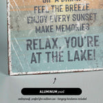 The image is a sign with text that reads "Sip a Drink, Feel the Breeze, Enjoy Every Sunset, Make Memories, Relax, You're at the Lake!" The sign is made of metal and has a black arrow pointing to it.