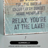 The image is a sign with text that reads "Sip a Drink, Feel the Breeze, Enjoy Every Sunset, Make Memories, Relax, You're at the Lake!" The sign is made of metal and has a black arrow pointing to it.