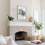 a cozy living room with a stone fireplace, a framed watercolor painting of purple hydrangeas hanging above it, and a white sofa. The room is decorated with potted plants and a woven basket, creating a warm and inviting atmosphere.