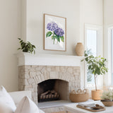 a cozy living room with a stone fireplace, a framed watercolor painting of purple hydrangeas hanging above it, and a white sofa. The room is decorated with potted plants and a woven basket, creating a warm and inviting atmosphere.