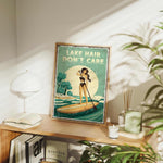 a framed poster of a woman paddleboarding on a lake, with the text "LAKESHAIR DON'T CARE" displayed prominently. The poster is placed on a wooden shelf, surrounded by various items such as books, plants, and a lamp.