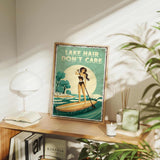 a framed poster of a woman paddleboarding on a lake, with the text "LAKESHAIR DON'T CARE" displayed prominently. The poster is placed on a wooden shelf, surrounded by various items such as books, plants, and a lamp.