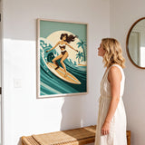 A woman in a white dress stands in a room, looking at a framed illustration of a woman surfing on a wave.
