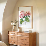 a wooden dresser with a framed painting of pink flowers hanging on the wall above it. The dresser has six drawers and a vase of white flowers on top.