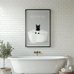 A framed poster with a black cat sitting in a white bathtub, surrounded by bubbles, is displayed on a white tiled wall above a white freestanding bathtub.