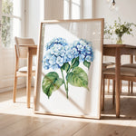 a framed painting of blue hydrangea flowers on a wooden table, with a vase of greenery and a wooden chair visible in the background.