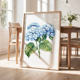 a framed painting of blue hydrangea flowers on a wooden table, with a vase of greenery and a wooden chair visible in the background.