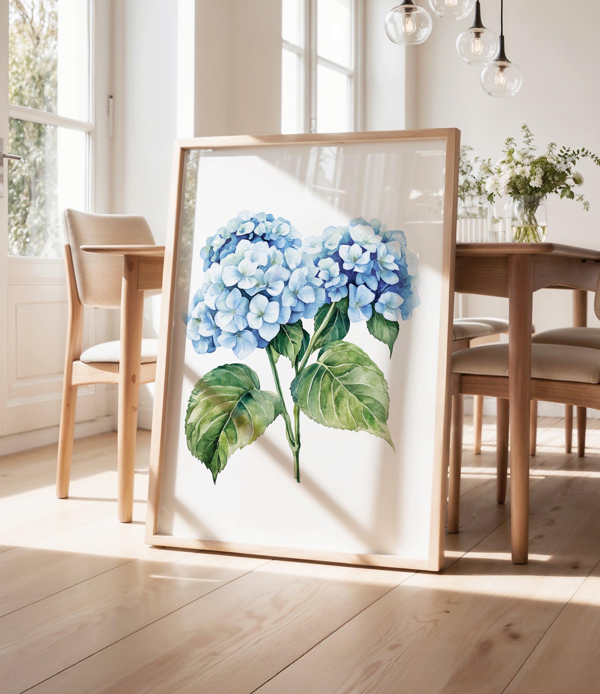 a framed painting of blue hydrangea flowers on a wooden table, with a vase of greenery and a wooden chair visible in the background.