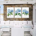 a cozy outdoor seating area with white wicker furniture and a stone wall backdrop. Three framed paintings of blue hydrangeas are displayed on the wall, creating a harmonious and visually appealing atmosphere.