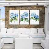 a cozy outdoor seating area with white wicker furniture and a stone wall backdrop. Three framed paintings of blue hydrangeas are displayed on the wall, creating a harmonious and visually appealing atmosphere.