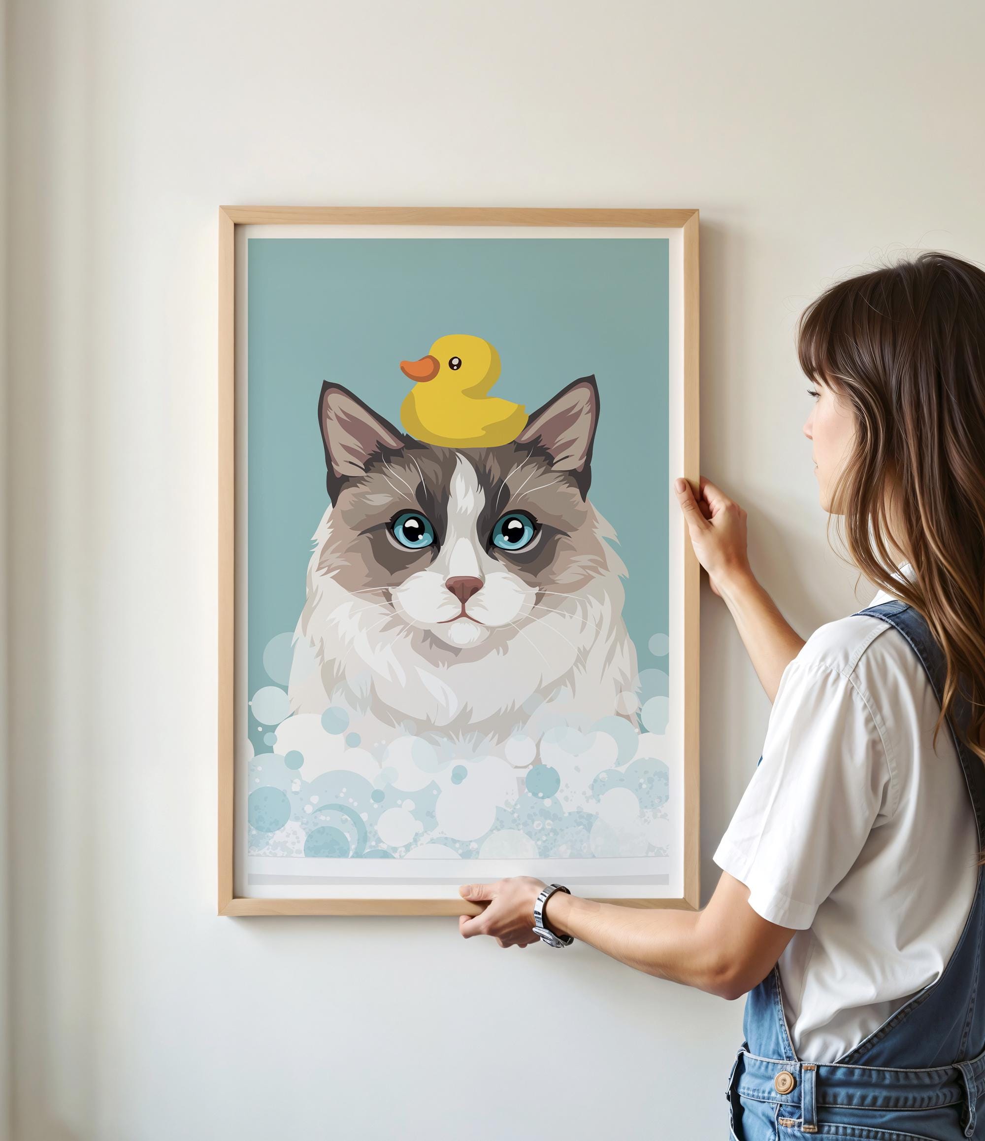 A woman is hanging a framed picture of a cat with a yellow rubber duck on its head.