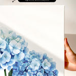 a hand holding a white canvas print of a watercolor painting of blue hydrangea flowers.