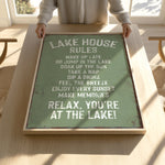 A person is holding up a framed sign with a list of rules for a lake house, including activities such as waking up late, jumping in the lake, taking a nap, drinking, feeling the breeze, and enjoying every sunset.