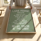 A person is holding up a framed sign with a list of rules for a lake house, including activities such as waking up late, jumping in the lake, taking a nap, drinking, feeling the breeze, and enjoying every sunset.
