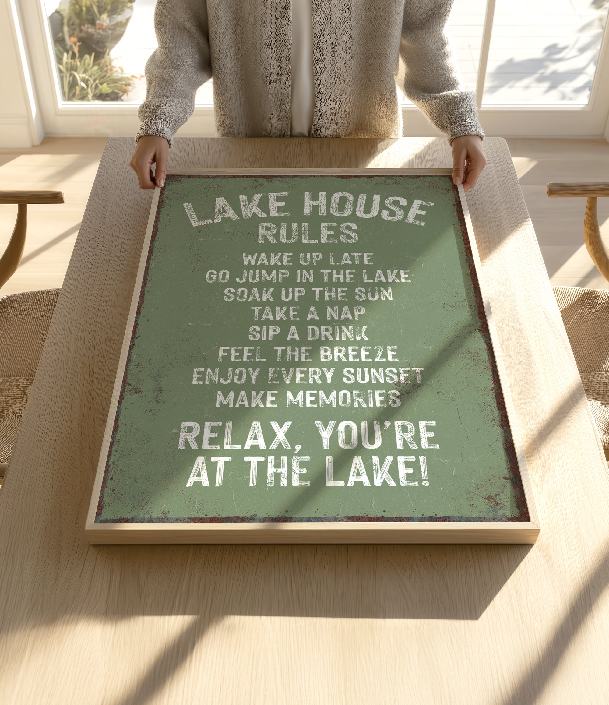 A person is holding up a framed sign with a list of rules for a lake house, including activities such as waking up late, jumping in the lake, taking a nap, drinking, feeling the breeze, and enjoying every sunset.