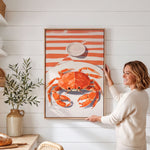 A woman is standing in a kitchen, holding up a framed picture of a crab on a white wall.