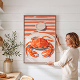 A woman is standing in a kitchen, holding up a framed picture of a crab on a white wall.