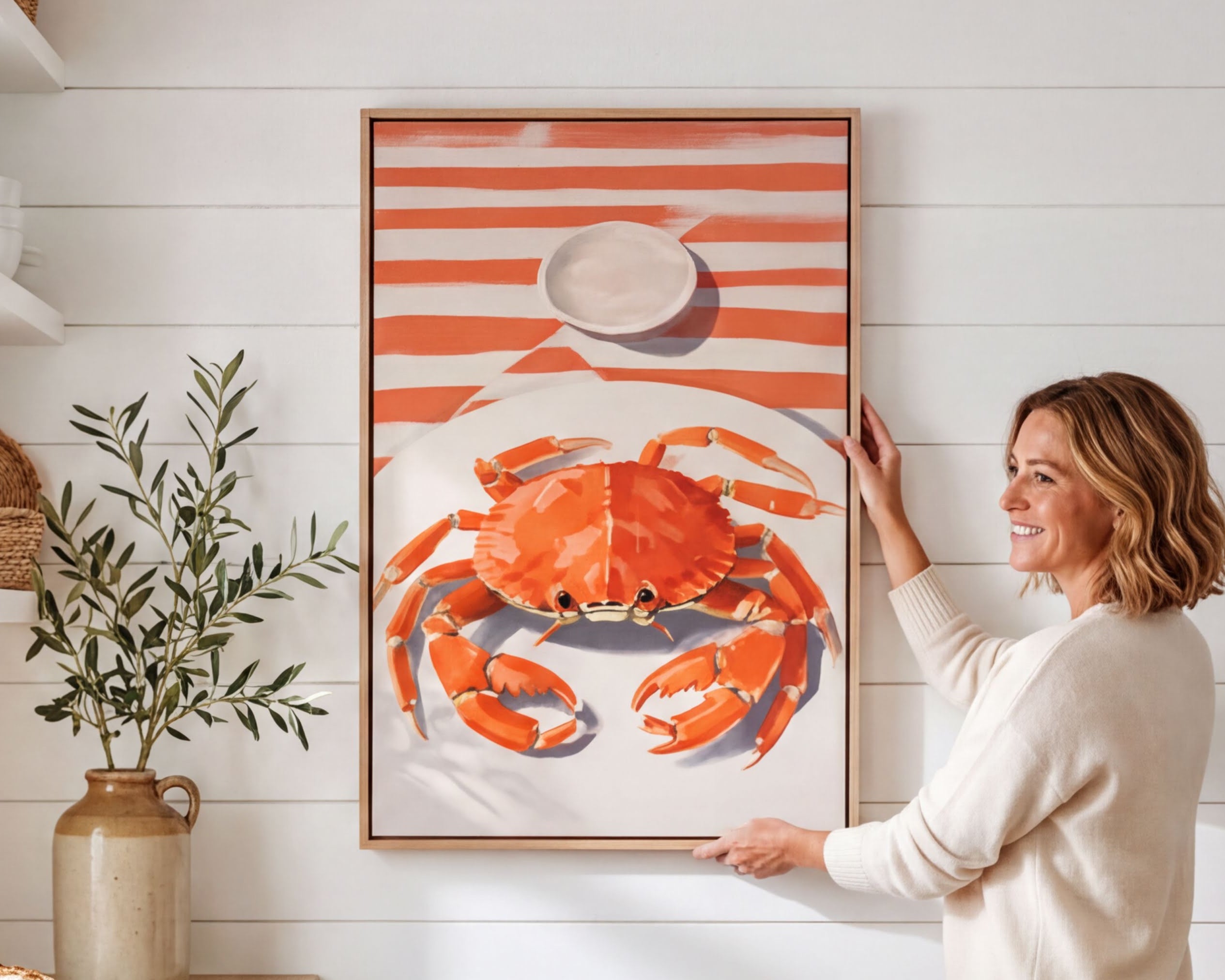 A woman is standing in a kitchen, holding up a framed picture of a crab on a white wall.
